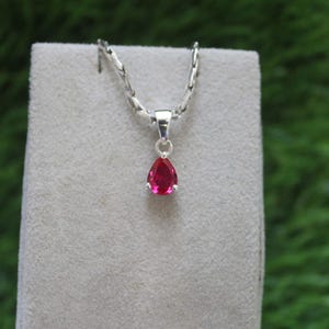 May include: A silver necklace with a pear-shaped, bright red gemstone pendant. The pendant is set in a silver bezel and hangs from a delicate silver chain. The necklace is displayed on a gray surface against a green background.