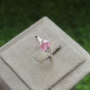 Könnte beinhalten: Ein silberner Ring mit einem rosafarbenen ovalen Edelstein, der von kleineren klaren Edelsteinen umgeben ist.