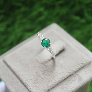 Peut inclure: Une bague en argent ornée d'un émeraude verte ronde et principale. La bague est ornée de petits pierres précieuses claires le long de la bande, créant un design délicat et élégant.