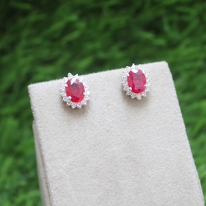 Peut inclure: Boucles d'oreilles à clous avec des pierres rouges ovales, entourées d'un halo de petites pierres claires. Les boucles d'oreilles sont serties dans un métal argenté et présentées sur une surface neutre, avec un fond vert flou.