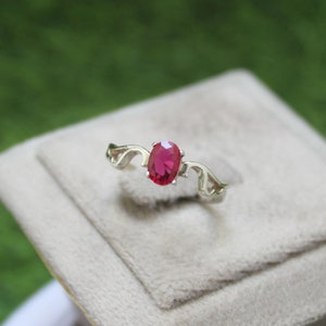 May include: A silver ring featuring a central oval-cut vibrant red gemstone held by four prongs. The band is crafted with open, wing-like metalwork extending from the setting on both sides, creating a distinct silhouette for this piece of jewelry.