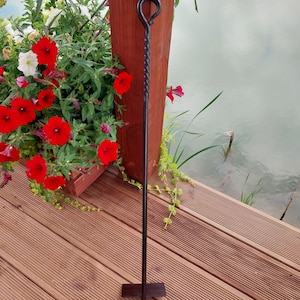 May include: A black wrought iron fire poker with a twisted handle and a loop at the top. The poker stands on a rectangular base. The background includes a wooden deck, a flower box with red and white flowers, and a body of water.