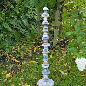 Op de afbeelding: Een hoge, verticale sculptuur bestaande uit talloze gladde, platte, veelkleurige stenen die in een enkele kolom zijn gestapeld. De stenen variëren in tinten grijs, wit, beige en zachtroze en rusten op een grote, ronde, gespikkelde grijze basissteen.