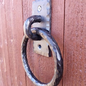 Può includere: Un battente per porta in metallo martellato con maniglia ad anello. Il battente è montato su una piastra rettangolare con superficie strutturata. L'anello è ovale e fissato alla piastra.