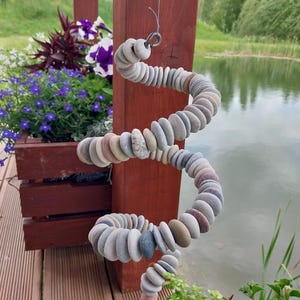 May include: A spiral-shaped hanging ornament constructed from numerous small, smooth, multi-colored river stones strung together on a wire. The stones vary in shades of gray, tan, and brown, forming a continuous, flexible, coiled structure.
