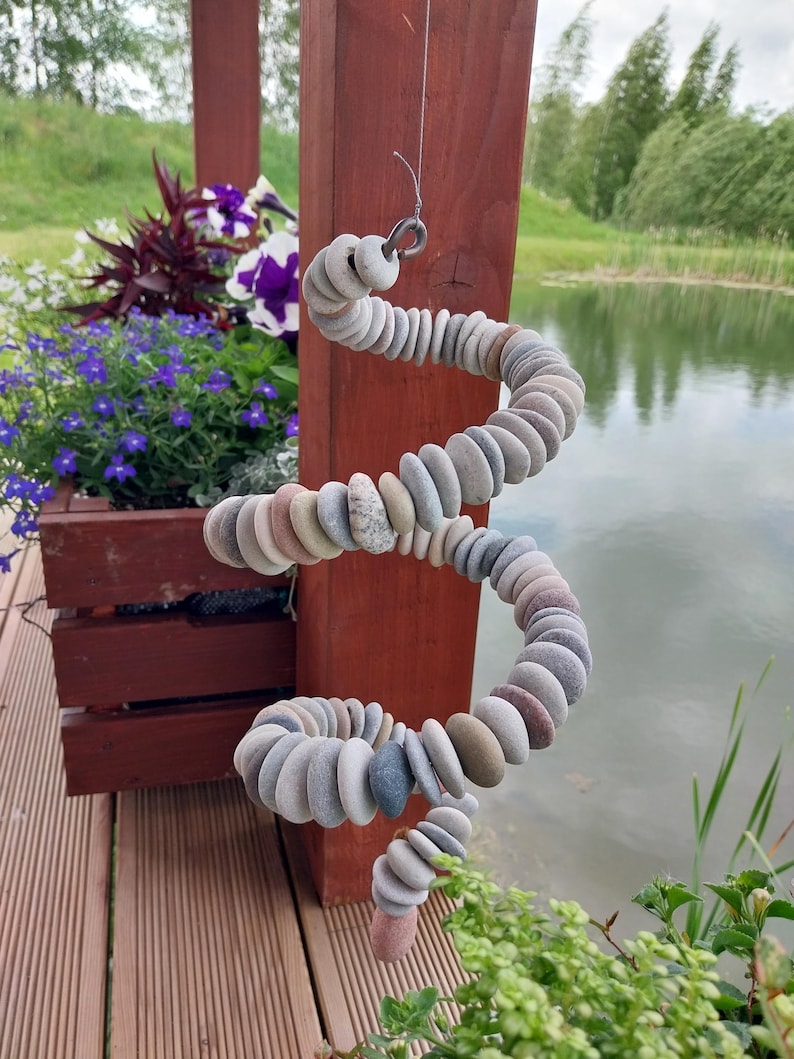 May include: A spiral-shaped hanging ornament constructed from numerous small, smooth, multi-colored river stones strung together on a wire. The stones vary in shades of gray, tan, and brown, forming a continuous, flexible, coiled structure.