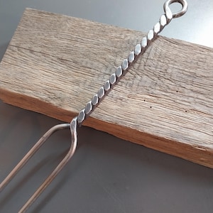 Könnte beinhalten: Ein handgeschmiedeter Metall-Kaminhaken mit gedrehtem Griff und einer Schlaufe oben. Das zweizinkige Ende dient zum Bewegen von Holzscheiten in einem Kamin oder einer Feuerstelle. Der Haken liegt auf einer verwitterten Holzoberfläche.