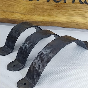 Könnte beinhalten: Drei schwarze, handgeschmiedete Metallgriffe mit strukturierter Oberfläche. Jeder Griff hat ein gebogenes Design mit Montageplatten an jedem Ende. Die Griffe werden vor einem hellen Hintergrund mit einem Holzschild im Hintergrund gezeigt.