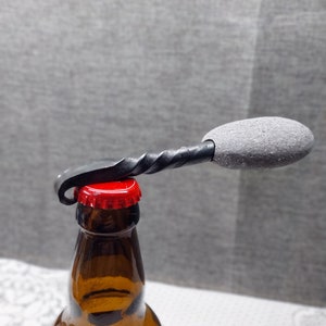 May include: A black metal bottle opener with a grey rubber handle. The opener is being used to open a brown glass bottle with a red cap.