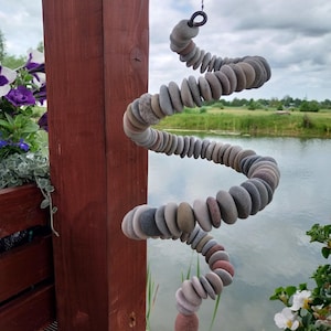 May include: A hanging spiral mobile constructed from numerous smooth, rounded river stones in various shades of gray, tan, and reddish-brown. The stones are strung together in a continuous, flexible coil that tapers to a single larger stone at the base.
