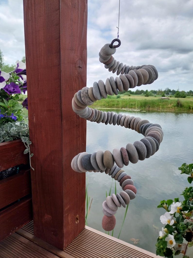 May include: A hanging spiral mobile constructed from numerous smooth, rounded river stones in various shades of gray, tan, and reddish-brown. The stones are strung together in a continuous, flexible coil that tapers to a single larger stone at the base.
