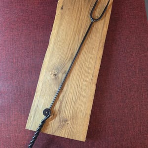 May include: A long, black, hand-forged metal roasting fork with a two-pronged head and a twisted handle featuring a spiral loop at the end. The fork is positioned diagonally across a rectangular piece of light brown wood with a natural live edge.