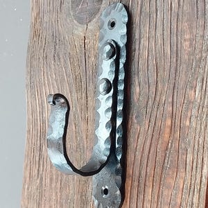 Puede incluir: Un gancho de metal forjado a mano con un acabado texturizado y martillado. El gancho está montado sobre una superficie de madera envejecida, mostrando una estética rústica. El diseño del gancho incluye remaches decorativos y una forma curva y funcional.