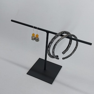 Può includere: Un espositore di gioielli in metallo nero con due braccialetti a cerchio argentati e un paio di orecchini pendenti. Gli orecchini hanno ganci argentati e perline gialle e grigie. I braccialetti hanno un motivo strutturato.