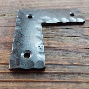 May include: A dark grey, L-shaped metal bracket with a hammered texture and three drilled holes. The edges have a decorative, scalloped design. The bracket is displayed on a wooden surface.