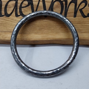 Op de afbeelding: Een zilverkleurige, gehamerde metalen ring met een getextureerd oppervlak. De ring lijkt handgemaakt. De achtergrond is een houten bord met het woord "Works" zichtbaar.