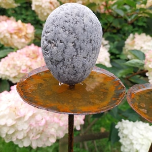 May include: A rustic bird bath featuring a grey stone perched on a rusty metal dish and stem. The background showcases pink and white hydrangea flowers and green leaves, creating a natural garden setting.