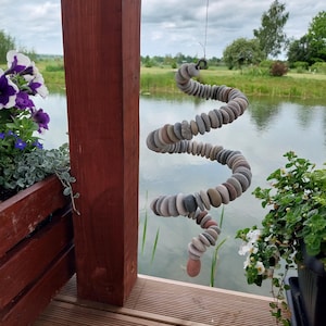 May include: A hanging spiral mobile constructed from numerous small, smooth, rounded river stones in various shades of gray, brown, and tan. The stones are strung together on a thin wire, forming a continuous, flexible, coiled shape suspended in air.