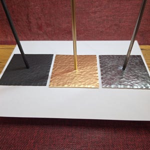 Pode incluir: Três suportes de exibição de metal com bases quadradas e hastes verticais. A base esquerda é preta fosca, a base central é dourada com textura martelada e a base direita é de metal prateado com uma textura martelada.