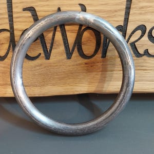 Puede incluir: Un anillo de metal plateado con una superficie texturizada. El anillo es circular y parece estar hecho de un material sólido. El fondo es una superficie de madera con la palabra "davworks" grabada.