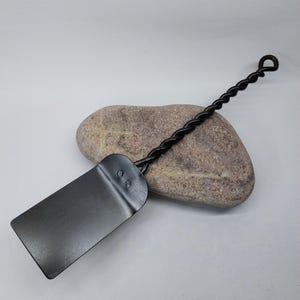 Puede incluir: Una espátula de metal negro con un mango retorcido. La espátula tiene un borde redondeado y dos pequeños remaches en la parte superior.