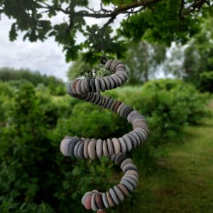 May include: A hanging spiral mobile constructed from numerous small, smooth, flat, multi-colored river stones in shades of gray, tan, and brown, suspended by a thin cord, creating a cascading, coiled shape that hangs vertically in the air.