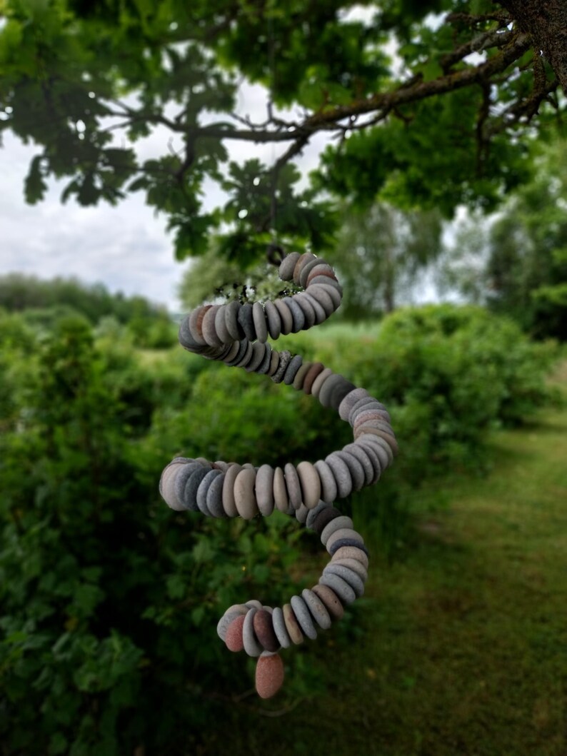 May include: A hanging spiral mobile constructed from numerous small, smooth, flat, multi-colored river stones in shades of gray, tan, and brown, suspended by a thin cord, creating a cascading, coiled shape that hangs vertically in the air.