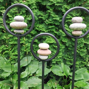 Peut inclure: Trois piquets de jardin en métal noir, chacun avec un cercle supérieur supportant des pierres lisses empilées. Les pierres varient en couleur du gris clair au beige. Les piquets sont plantés dans un jardin verdoyant, avec un fond de feuillage persistant dense.