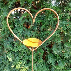May include: A rustic, heart-shaped metal garden stake with a small, round dish. The stake is made of weathered, rusty metal and is set against a backdrop of green foliage. The heart measures approximately 12 inches wide.