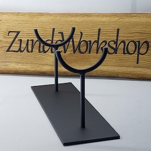 Puede incluir: Soporte de metal negro con dos brazos curvos para exhibir artículos. El soporte tiene una base rectangular y está hecho de metal. El soporte está sobre una superficie blanca. El texto "Zunda Workshop" es visible en un letrero de madera detrás del soporte.