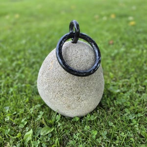 May include: A black metal ring with a hammered, textured surface. The ring is circular and features a smaller, integrated loop at the top, creating a pendant-like structure. The metal has a dark, glossy finish that reflects light across its surface.