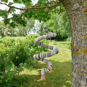 May include: A hanging spiral mobile constructed from numerous small, smooth, rounded stones in shades of gray, tan, and muted pink. The stones are strung together in a continuous, twisting coil shape, suspended by a thin cord from a tree branch.