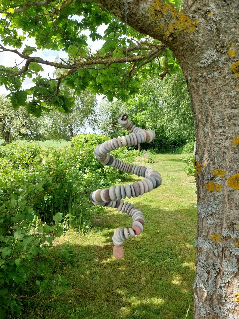 May include: A hanging spiral mobile constructed from numerous small, smooth, rounded stones in shades of gray, tan, and muted pink. The stones are strung together in a continuous, twisting coil shape, suspended by a thin cord from a tree branch.