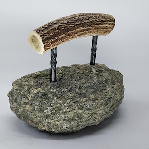 Op de afbeelding: Een kleine, groene steen met twee gedraaide metalen staven die eruit steken. Een bruin geweistuk is bovenop de staven bevestigd.