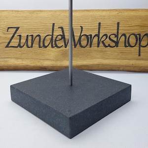 Op de afbeelding: Een donkergrijze vierkante displaystandaard met een verticale metalen staaf. De standaard is gemaakt van een solide, getextureerd materiaal. De achtergrond toont een houten bord met de tekst "Zunde Workshop" in zwart.