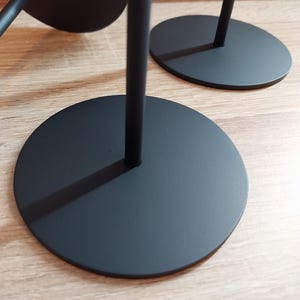 Puede incluir: Primer plano de soportes decorativos de metal negro. Cada soporte tiene una base circular y un tallo vertical y delgado. Los soportes están colocados sobre una superficie de madera de color claro, con un enfoque en el diseño minimalista y el contraste.