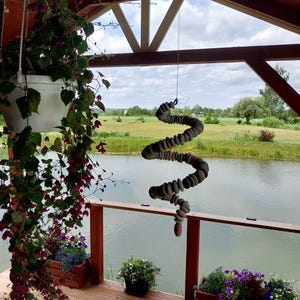 May include: A hanging mobile sculpture constructed from a series of smooth, rounded grey stones threaded together in a spiral shape, suspended by a thin wire from the wooden ceiling of an outdoor porch overlooking a pond and green landscape.