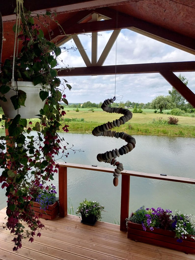 May include: A hanging mobile sculpture constructed from a series of smooth, rounded grey stones threaded together in a spiral shape, suspended by a thin wire from the wooden ceiling of an outdoor porch overlooking a pond and green landscape.