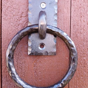 Könnte beinhalten: Ein handgeschmiedeter Türklopfer aus dunklem Metall mit gehammerter Textur. Der Klopfer hat einen großen, kreisförmigen Ring, der an einer rechteckigen Rückplatte mit einem kleinen, kreisförmigen Emblem befestigt ist. Der Klopfer ist an einer rotbraunen Holztür angebracht.