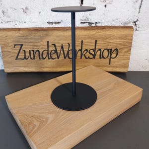 Op de afbeelding: Een zwarte metalen displaystandaard met een cirkelvormige boven- en onderkant, verbonden door een slanke steel. Het staat op een lichtgekleurd houten blok. Het houten blok heeft de tekst "ZundeWorkshop" gegraveerd.