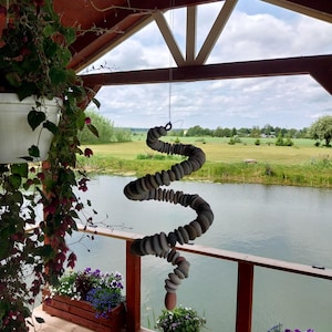 May include: A hanging mobile sculpture constructed from numerous smooth, rounded gray and tan stones strung together in a spiral formation. The stones are arranged in a vertical, twisting column suspended by a thin wire from a wooden structure.