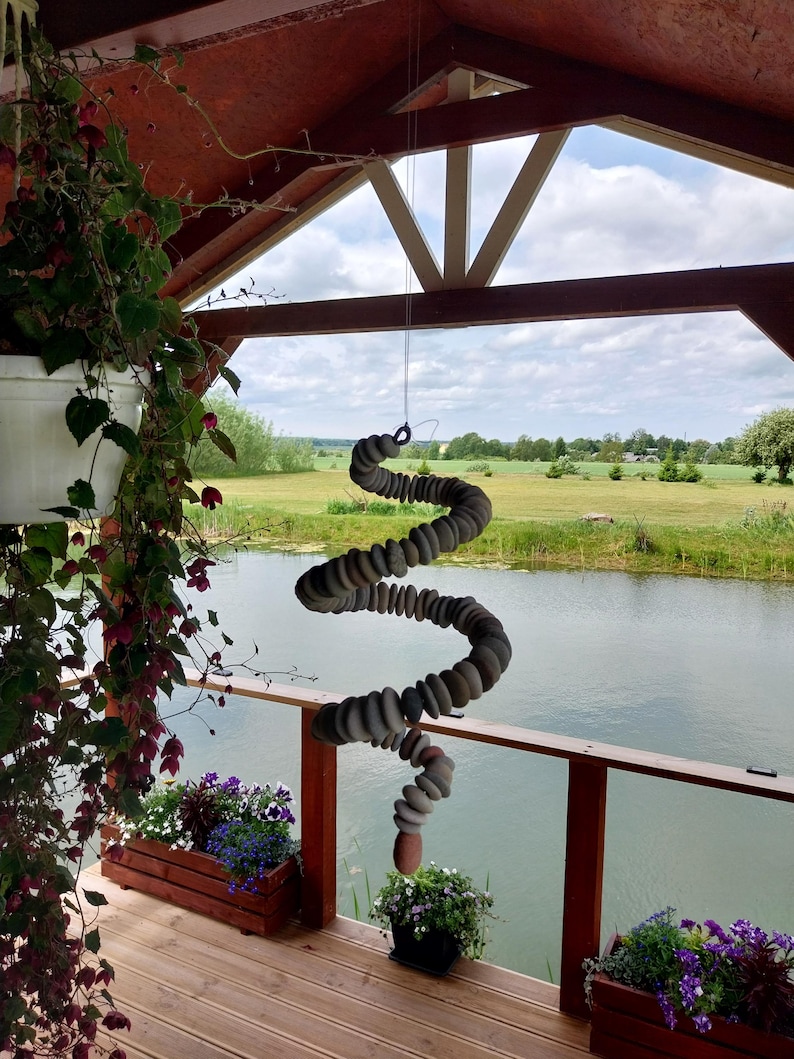 May include: A hanging mobile sculpture constructed from numerous smooth, rounded gray and tan stones strung together in a spiral formation. The stones are arranged in a vertical, twisting column suspended by a thin wire from a wooden structure.