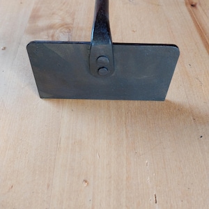 Puede incluir: Una azada de jardín de metal oscuro con una hoja rectangular y un mango largo. La hoja está unida al mango con dos remaches. La herramienta está diseñada para tareas de jardinería y paisajismo.