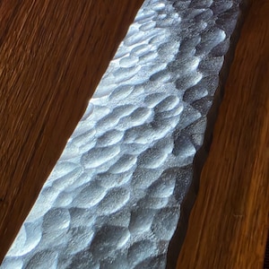 Könnte beinhalten: Eine lange, rechteckige Metallstange mit einer ausgeprägten gehämmerten, genoppten Textur auf der Oberfläche. Das Metall hat ein dunkles, anthrazitfarbenes Finish, wobei sich das Licht an den Kanten der Vertiefungen bricht, auf Holz.
