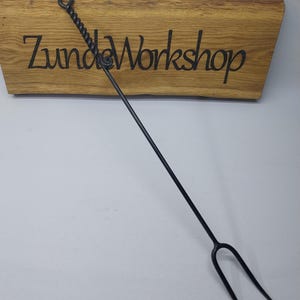 Op de afbeelding: Een zwarte metalen braadvork met een gedraaide handgreep en een lusvormig uiteinde. De vork heeft twee tanden en wordt getoond tegen een houten bord met de tekst "Zunda Workshop".
