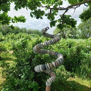 May include: A hanging spiral mobile constructed from numerous smooth, rounded, multi-colored river stones in shades of gray, tan, and brown. The stones are strung together in a continuous, twisting vertical column suspended from a tree branch.