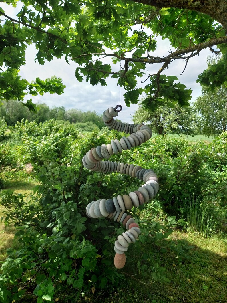 May include: A hanging spiral mobile constructed from numerous smooth, rounded, multi-colored river stones in shades of gray, tan, and brown. The stones are strung together in a continuous, twisting vertical column suspended from a tree branch.