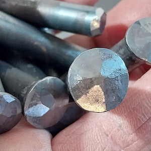 May include: Close-up of several hand-forged, dark metal nails. The nail heads have a faceted, geometric design, reflecting light with a metallic sheen. The cylindrical shafts are also visible, with a textured surface. The nails are held in a hand.