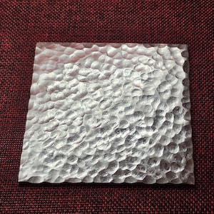 Puede incluir: Un posavasos cuadrado de metal de color plateado que presenta una textura martillada distintiva en toda su superficie superior. El acabado es reflectante y crea un patrón de pequeñas hendiduras redondeadas que captan la luz, dando al objeto una apariencia texturizada.