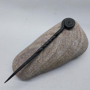 May include: A black metal hair pin with a spiral design at the top and a twisted shaft. The pin is resting on a grey stone, showcasing its handcrafted details. This unique accessory adds a touch of artistry to any hairstyle.